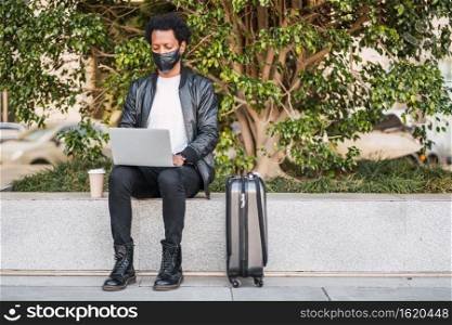 Portrait of afro tourist man using his laptop and wearing protective mask while sitting outdoors. Tourism concept.