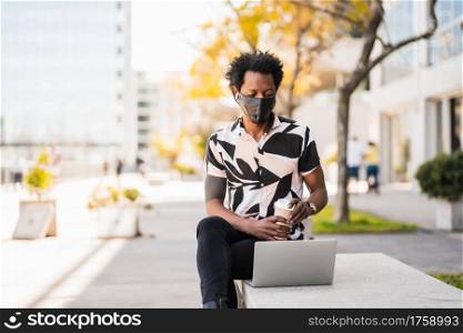 Portrait of afro tourist man using his laptop and wearing protective mask while sitting outdoors. New normal lifestyle concept.