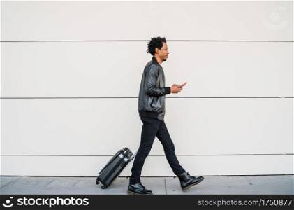 Portrait of afro tourist man using his mobile phone and carrying suitcase while walking outdoors on the street. Tourism concept.