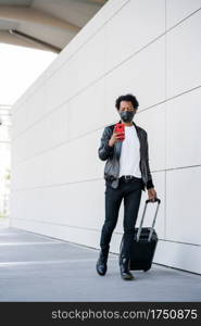 Portrait of afro tourist man using his mobile phone and carrying suitcase while walking outdoors. Tourism concept. New normal lifestyle concept.