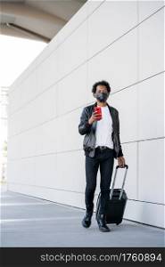 Portrait of afro tourist man using his mobile phone and carrying suitcase while walking outdoors. Tourism concept. New normal lifestyle concept.