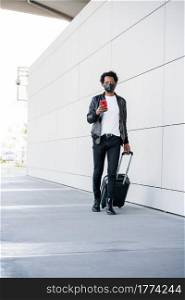 Portrait of afro tourist man using his mobile phone and carrying suitcase while walking outdoors. Tourism concept. New normal lifestyle concept.. Tourist man using his mobile phone and carrying suitcase outdoors.