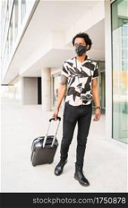 Portrait of afro tourist man wearing protective mask and carrying suitcase while walking outdoors on the street. Tourism concept.