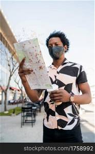 Portrait of afro tourist man wearing protective mask and looking for directions on map while walking outdoors on the street. Tourism concept.