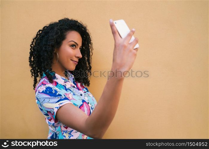 Portrait of afro woman taking selfies with her mophile phone against yellow wall. Technology concept.