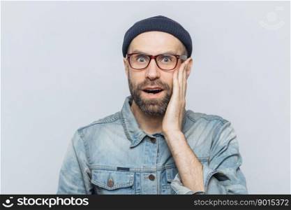 Portrait of amazed bearded excited male looks with surprisment in camera, recieves unexpected news, dressed in fashionable clothing, has attractive look, isolated over white studio background