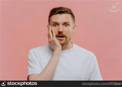 Portrait of amazed unshaven redhead man expressing astonishment while looking at camera with surprised shocked face expression, wondering while standing isolated over pastel pink studio background.. Portrait of surprised man looking at camera with shocked face expression
