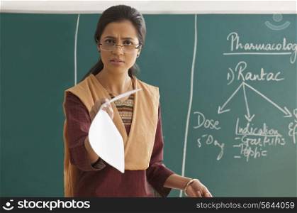 Portrait of an angry female teacher holding paper against chalkboard