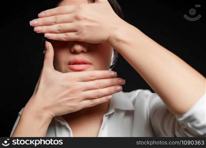 portrait of an Asian brunette woman in a white shirt covering her face with her hand on a black background