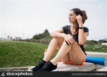 Portrait of an athletic woman listening to music on a break from training while sitting in park. Sport and health lifestyle concept.