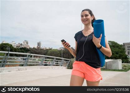 Portrait of an athletic woman walking on the street holding a training mat while listening to music. Sport and lifestyle concept.