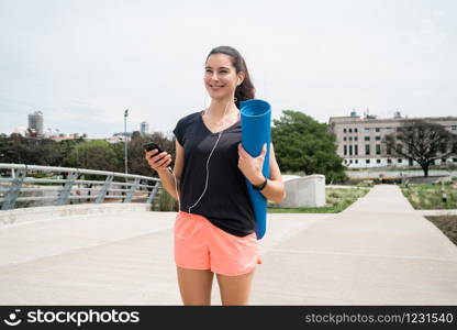 Portrait of an athletic woman walking on the street holding a training mat while listening to music. Sport and lifestyle concept.