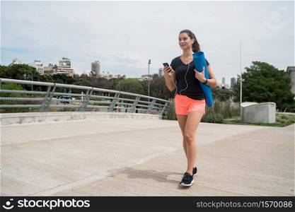 Portrait of an athletic woman walking on the street holding a training mat while listening to music. Sport and lifestyle concept.