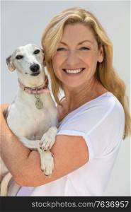Portrait of an attractive middle aged blonde woman smiling with perfect teeth holding her pet whippet dog