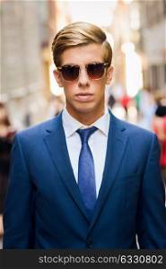 Portrait of an attractive young businessman in urban background wearing blue suit, sunglasses and necktie. Blonde hair