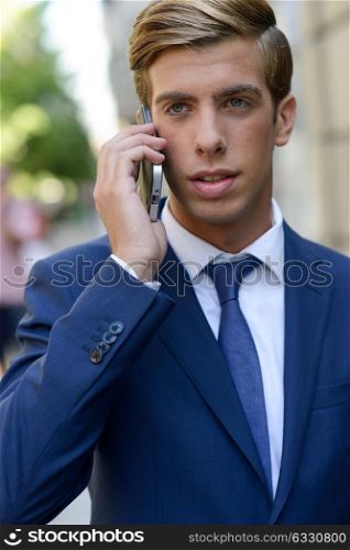 Portrait of an attractive young businessman on the phone in urban background, wearing blue suit and tie. Blonde hair