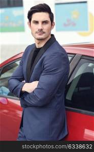 Portrait of an attractive young businessman outdoors, wearing modern suit, standing near a red car.