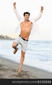 Portrait of an attractive young man jumping on a tropical beach. Handsome guy wearing white shirt and striped shorts. Male with beautiful body.