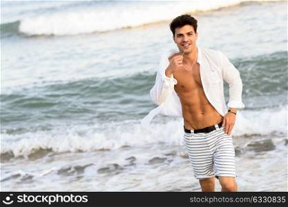 Portrait of an attractive young man on a tropical beach. Handsome guy wearing white shirt and striped shorts. Male with beautiful body.