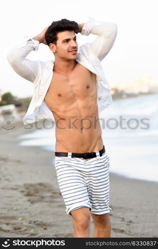 Portrait of an attractive young man on a tropical beach. Handsome guy wearing white shirt and striped shorts. Male with beautiful body.