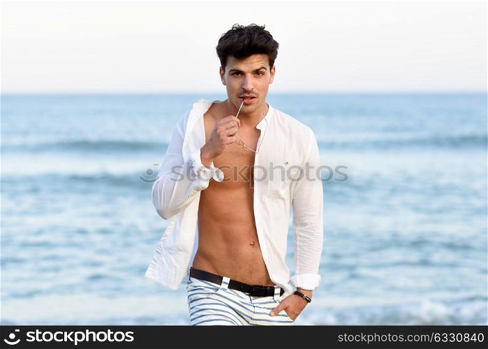 Portrait of an attractive young man on a tropical beach. Handsome guy wearing white shirt and striped shorts. Male with beautiful body and eyeglasses