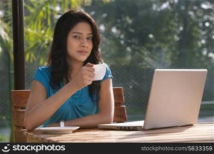 Portrait of an attractive young woman having tea while sitting by table with laptop
