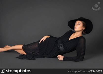 Portrait of an elegant young white woman with wavy hair and beautiful makeup laying by herself on the floor inside a studio with a grey background wearing a black dress with a belt and hat.