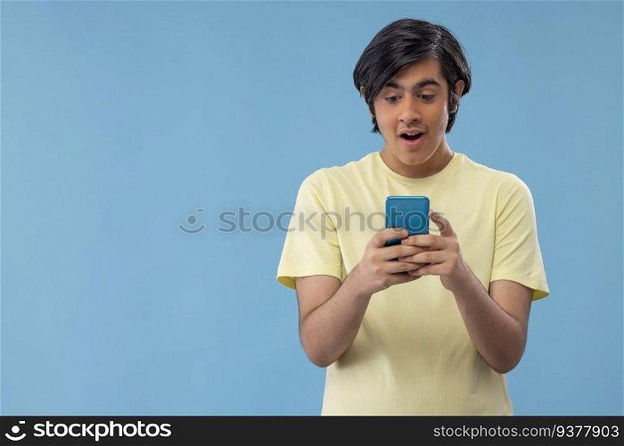 Portrait of an excited teenage boy using Smartphone while standing against blue background