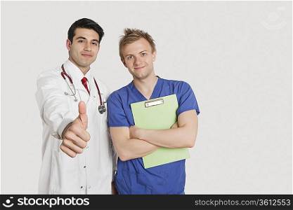 Portrait of an Indian doctor gesturing thumbs up while standing with male nurse over light gray background