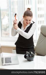 Portrait of angry woman at office hitting piggy bank with hammer