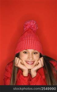 Portrait of Asian-American teen girl wearing winter hat resting head on hands against red background.