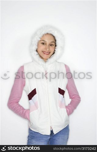 Portrait of Asian-American teen girl with hands in coat pockets and furry hood on head smiling against white background.