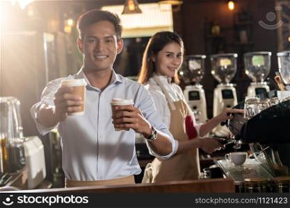 Portrait of asian barista hold take away coffee cup and serving to customer in cafe with other barista working in background
