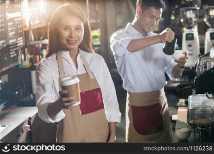 Portrait of asian barista hold take away coffee cup and serving to customer in cafe with other barista working in background