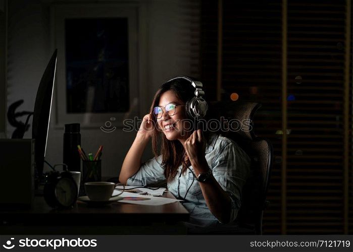 Portrait of Asian Businesswoman listening the music via headphone and smart mobile phone with glad action on the table in workplace at late time, Work hard and too late concept