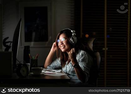 Portrait of Asian Businesswoman listening the music via headphone and smart mobile phone with glad action on the table in workplace at late time, Work hard and too late concept