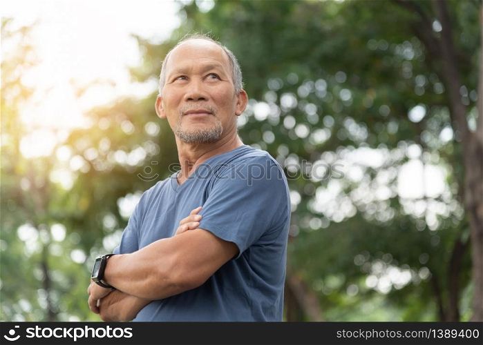 Portrait of Asian Elderly man with arms crossed relaxing over nature background. Happy confident Senior person in blue shirt standing at the park. Positive emotion, Carefree, Looking away, Health care, Lifestyle, Retirement.