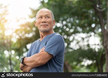 Portrait of Asian Elderly man with arms crossed relaxing over nature background. Happy confident Senior person in blue shirt standing at the park. Positive emotion, Carefree, Looking away, Health care, Lifestyle, Retirement.