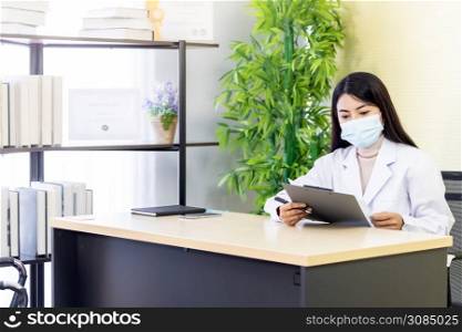 Portrait of asian female doctor wear protective face mask sit in her office room in hospital clinic and reading on patience file before checking patience. New Normal Health care Concept.