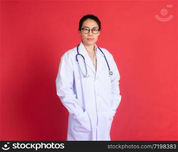 Portrait of Asian female doctor with stethoscope and glasses isolated on red background standing with cheerful gesture wearing doctor uniform with smiling face. Health insurance & physician concept.