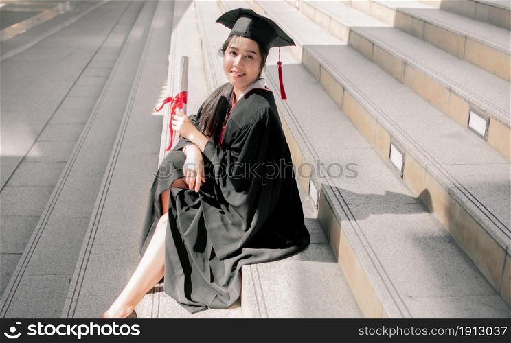 Portrait of Asian female student wearing graduated uniform and holding certification while sitting on stairs. Education Concept.