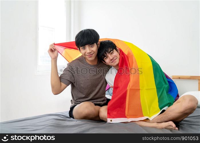 Portrait of Asian gay couple smiling on face, looking at camera, covering body by rainbow flag and sitting on the bed. Asian homosexsual man celebrating together with pride flag in bedroom.