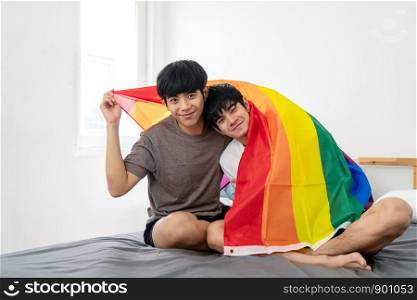 Portrait of Asian gay couple smiling on face, looking at camera, covering body by rainbow flag and sitting on the bed. Asian homosexsual man celebrating together with pride flag in bedroom.