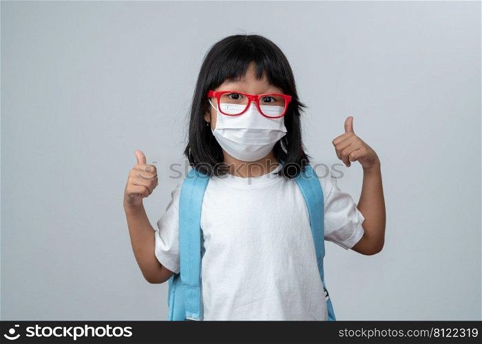 Portrait of Asian girl kid with protective face mask and school backpack and thumbs up, ready for new school year. Concept of kid going back to school and new normal lifestyle