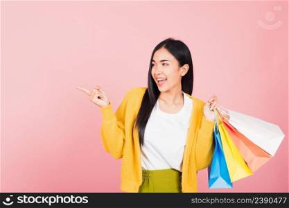 Portrait of Asian happy beautiful young woman shopper smiling standing excited holding online shopping bags multicolor in summer pointing finger to copy space, studio shot isolated on pink background