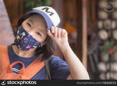 Portrait of Asian LGBT woman in fabric face mask with cap looking at camera with self confidence in her eyes on blurred vintage outdoor market background, diversity of people concept