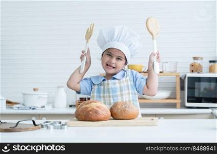 Portrait of Asian litt≤cute kid girl wear chef hat holding wooden fork and spoon, smi≤at camera with fresh loaf of bread in kitchen at home or cooking class at school. Child education concept