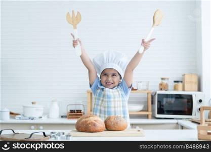 Portrait of Asian little cute kid girl wear chef hat holding wooden fork and spoon, raise hands up, with fresh loaf of bread in kitchen at home or cooking class at school. Child education concept