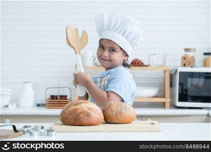 Portrait of Asian little cute kid girl wear chef hat holding wooden fork and spoon, smile at camera with fresh loaf of bread in kitchen at home or cooking class at school. Child education concept