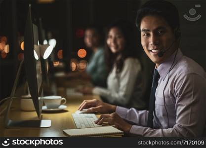 Portrait of Asian Male customer care service with businesswoman smiling and working hard late in night shift at office,call center department,worker and overtime,team work with colleagues for success